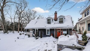 A snow-covered house with a brick chimney, a red door, and a small porch area.