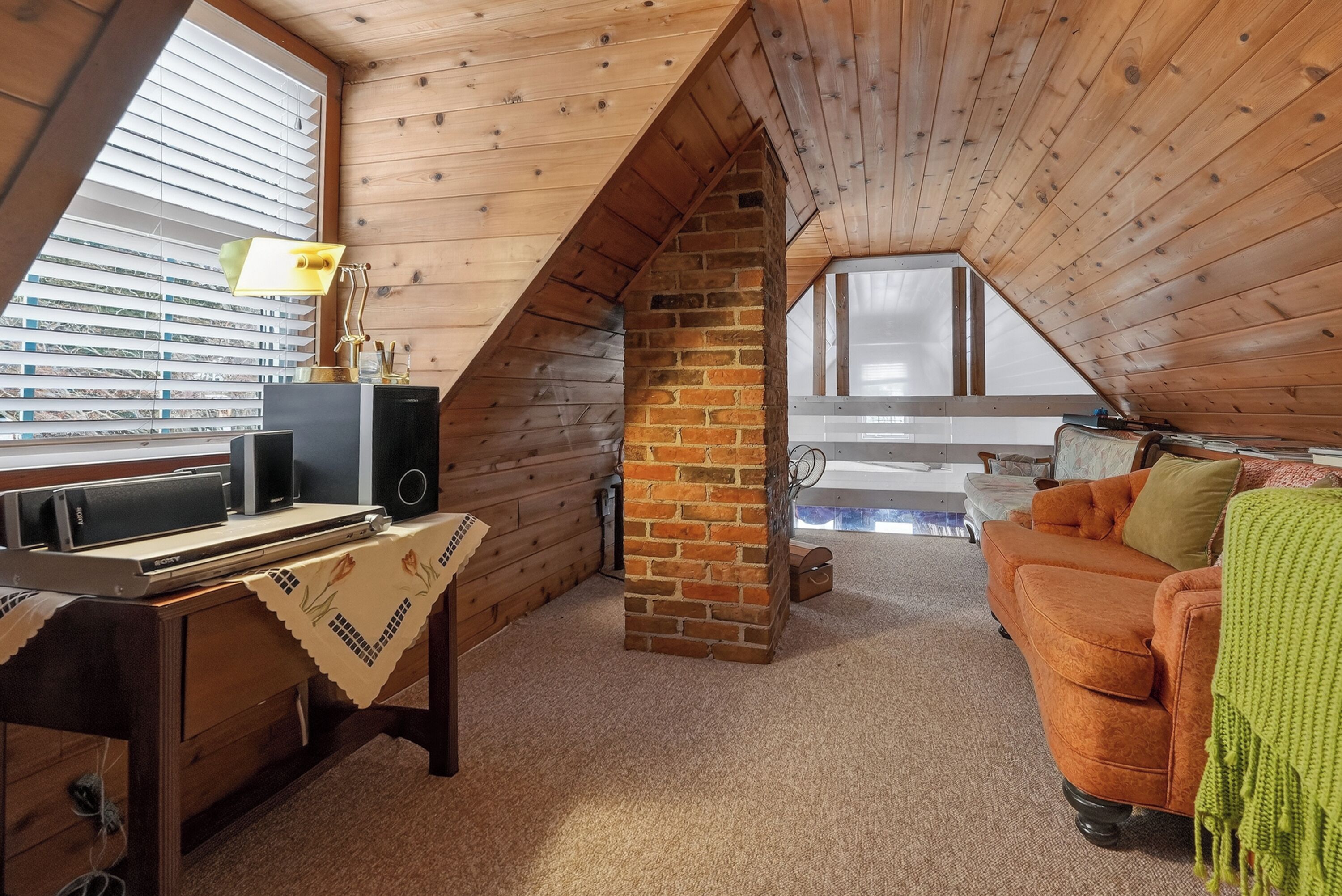 Una habitación cálida en el desván, con techo inclinado, chimenea de ladrillo, un escritorio de madera con lámpara, un sofá y una ventana con persianas.
