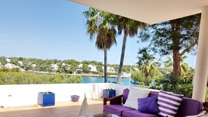 A terrace with a glass table, purple sofa, and a view of a coastal area with buildings and trees. - VILLA SA CALETA:  6 beds, 5 baths  Front line Villa In Es Forti, Cala d´Or (Santanyí)