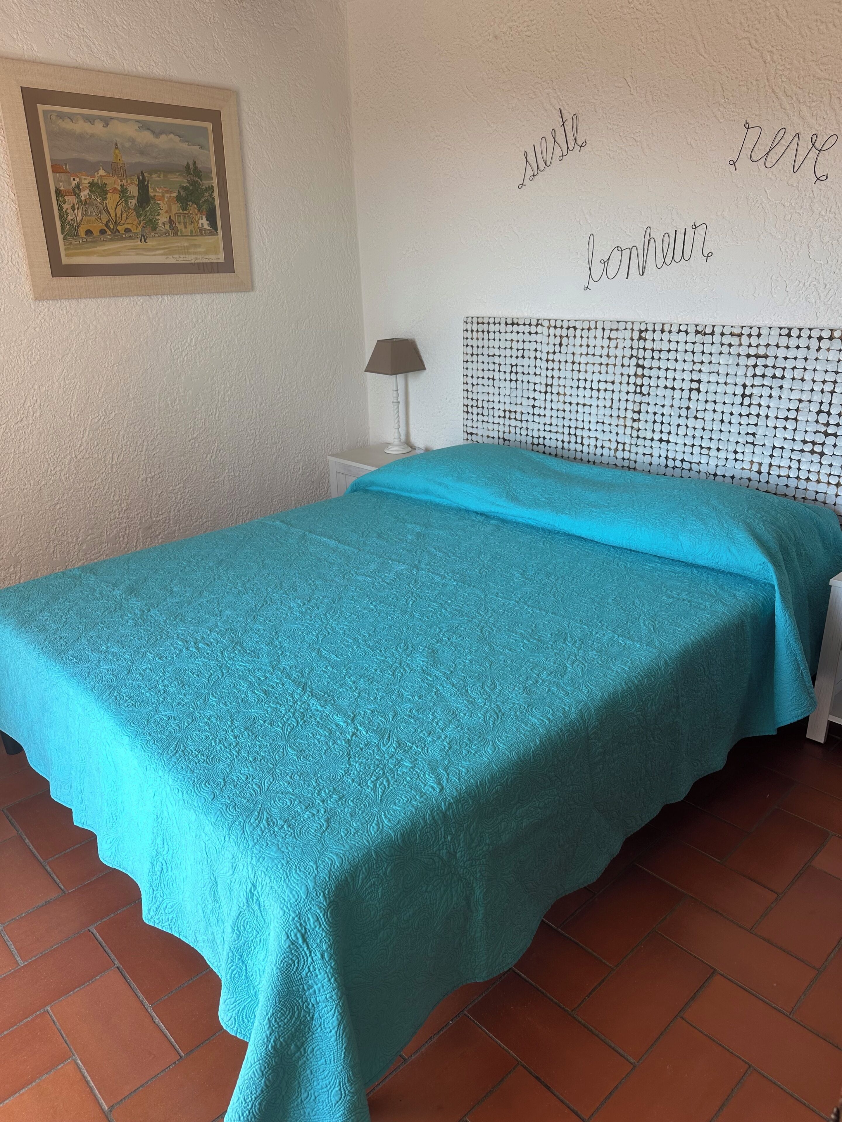 A room with a bed, a nightstand, a lamp, and a wall with a framed painting and text.