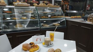 Un café con una vitrina llena de pasteles y pan, una mesa con una bebida y pasteles, y un cartel con el menú.