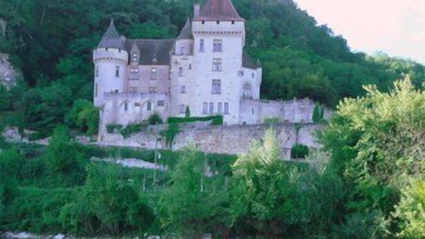 A castle-like building with multiple towers, situated on a hillside overlooking a calm river.