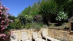 A stone wall with lounge chairs and a table, surrounded by flowering plants and trees.