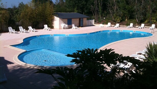 Piscina com espreguiçadeiras e um pequeno edifício, cercada por árvores.