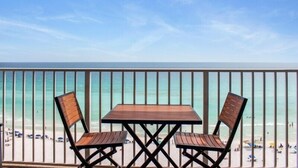 Ein Balkon mit Holztisch und zwei Stühlen, der Blick geht auf einen Strand mit Menschen und Sonnenschirmen.