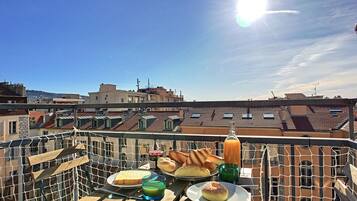 A rooftop terrace with a table set for breakfast, including bread, pastries, and a glass of juice.