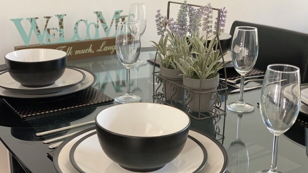 Een eettafel gedekt met zwarte kommen, witte borden en wijnglazen, met een potplant en een decoratief bordje.