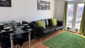 Een moderne woonkamer met een eetgedeelte, met een leren zwarte bank, een glazen eettafel en een stadsgezicht door een groot raam.