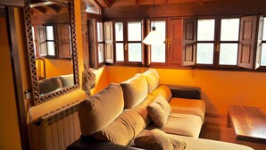Een gezellige kamer met een houten plafond, een bank met meerdere kussens, een houten tafel en een raam met houten luiken.