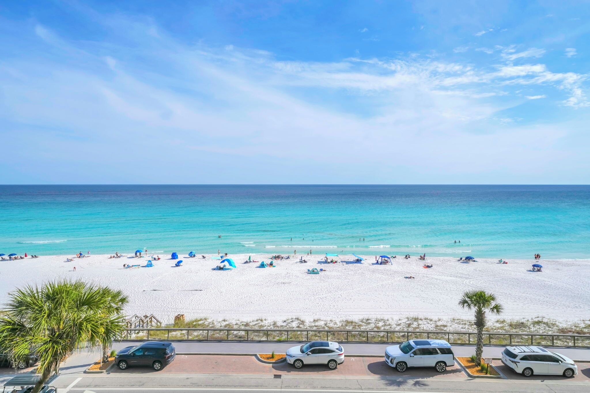 A beach with white sand, clear blue water, and a few parked cars.