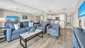 Una sala moderna con un sofá, sillones, una mesa de centro y un televisor. Se ve un ventilador de techo, una zona de comedor con sillas y una cocina al fondo.
