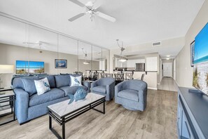 A modern living room with a sofa, armchairs, a coffee table, and a TV. There is a ceiling fan, a dining area with chairs, and a kitchen in the background.
