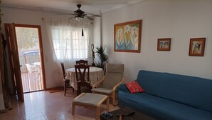 A living room with a blue sofa, a wooden coffee table, a dining table with chairs, and a ceiling fan.