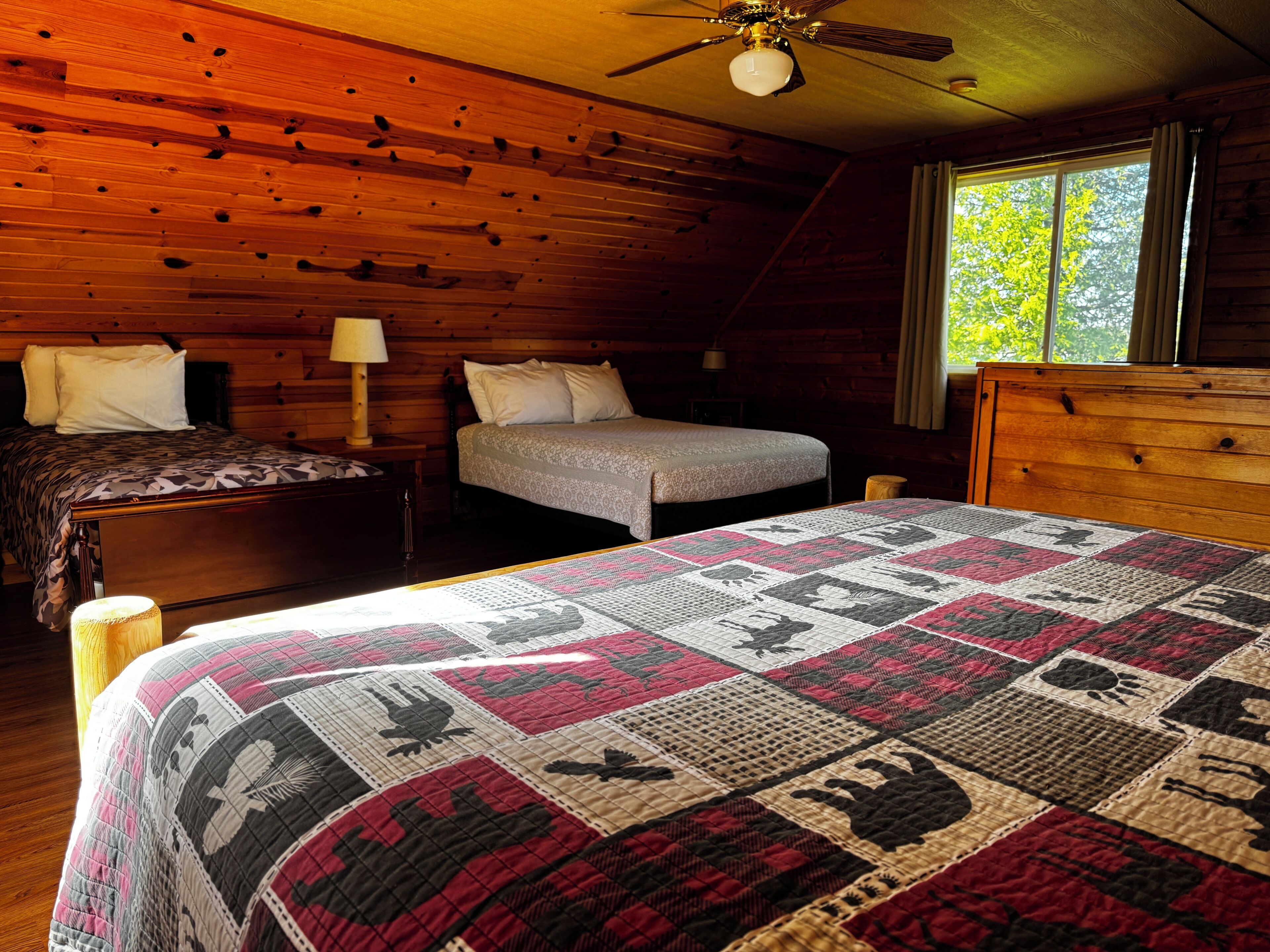 A cabin-style bedroom with a wooden ceiling, a bed with a quilt, a nightstand, a dresser, and a window with a view of trees.