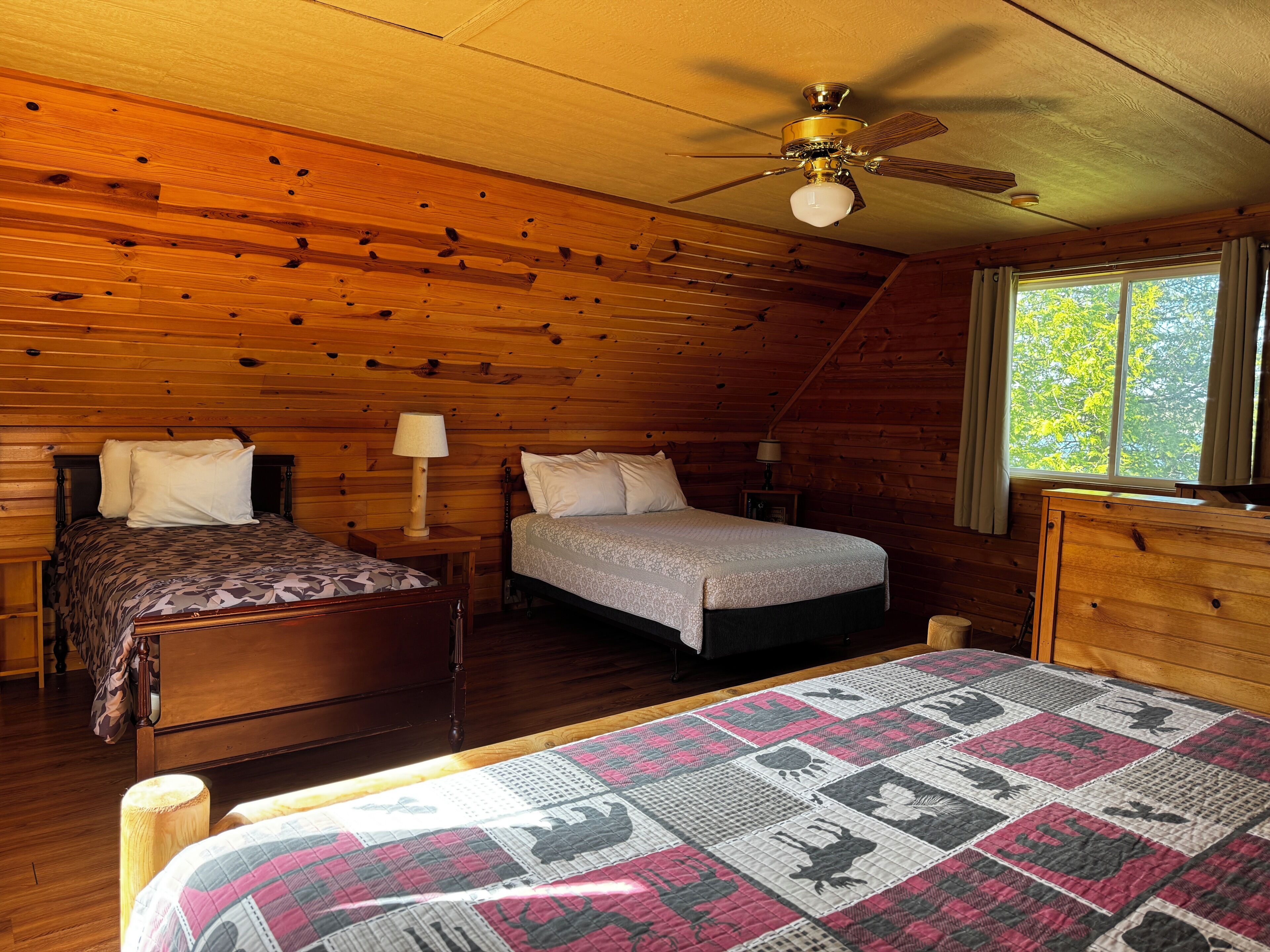 A cabin-style room with two wooden beds, a ceiling fan, and a large window.