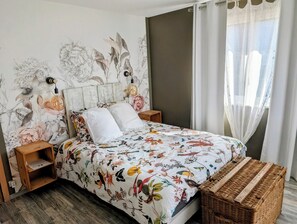 A bedroom with a floral bedspread, a wooden headboard, bedside tables, a wicker basket, and a wall with a floral mural. - Lovely, comfortable detached house near Colmar and vineyards (Sundhoffen)