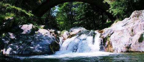 A stone bridge over a river with a waterfall, surrounded by rocks and greenery.
