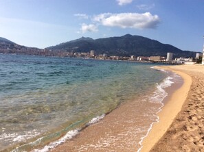 Uma praia com água cristalina, areal dourado e paisagem urbana ao fundo.