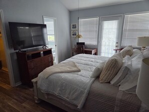 Une chambre à coucher avec un lit, un téléviseur posé sur une commode en bois, un bureau avec une chaise et une fenêtre avec des stores.