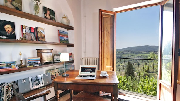 Een kamer met een houten bureau, een laptop, een kopje en uitzicht op een groen landschap door een balkondeur.