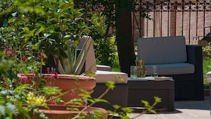 Een terras met een rieten bank en een bijzettafeltje, omgeven door potplanten en een bakstenen muur.