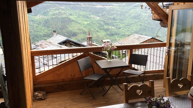 Een houten terras met een tafel en stoelen, een vaas met bloemen en uitzicht op een bebost dal met gebouwen.