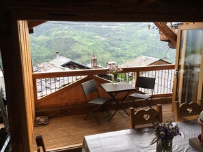Eine Holzterrasse mit Tisch und Stühlen, eine Vase mit Blumen und Blick auf ein bewaldetes Tal mit Gebäuden.
