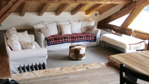 Een knus zolderkamertje met een houten plafond, een zithoek, een rond poefje en een kleine tafel.