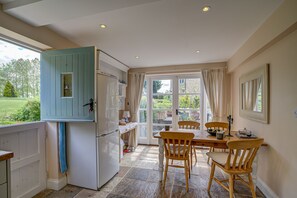 A kitchen with a blue cabinet, a dining table with chairs, and a view of the garden through the door. - Spring Hill Cottage | Oxfordshire Holiday Let | 2 Beds & Sleeps 4 (Begbroke)