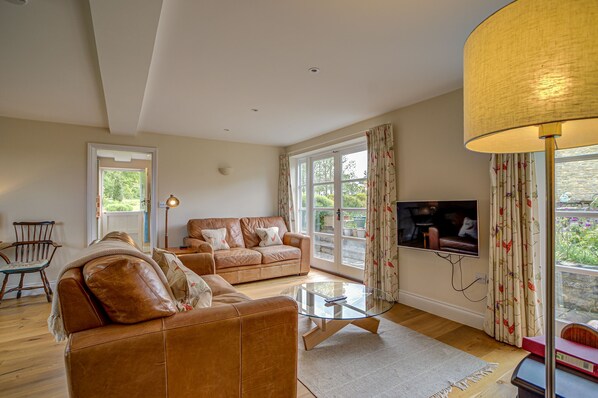 A living room with a brown leather sofa, a glass coffee table, and a television on the wall. - Spring Hill Cottage | Oxfordshire Holiday Let | 2 Beds & Sleeps 4 (Begbroke)