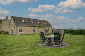 A stone house with a sloped roof and skylights, a grassy lawn, and a dining area with metal chairs and a table. - Spring Hill Cottage | Oxfordshire Holiday Let | 2 Beds & Sleeps 4 (Begbroke)