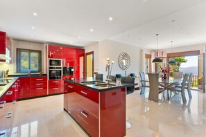 A modern kitchen with red cabinets, a central island, and a dining area with a glass table and chairs. - Ultra-Luxury Villa with Private Pool, Art Deco Cinema & 1950s Diner– Sleeps 6 (Mijas)