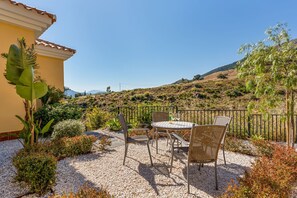 Eine Terrasse mit Tisch und Stühlen, umgeben von Kies und Pflanzen, mit einem Zaun und Hügeln im Hintergrund.