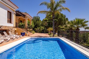 Ein Schwimmbecken mit Liegestühlen, einer überdachten Terrasse und Blick auf Bäume sowie ein Gebäude.