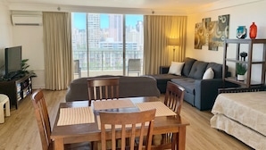 Un salon moderne comprenant un coin repas, un ensemble de canapés, un téléviseur et un balcon avec vue sur la ville.