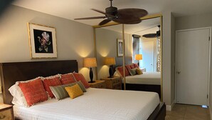A bedroom with a large bed, a ceiling fan, a mirror, and a door. - Affordable Paradise  (Kihei)