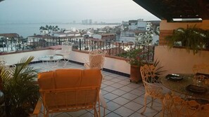 Eine Terrasse mit Blick auf eine Stadtlandschaft und Wasser. Es gibt Stühle und einen Tisch mit einer Pflanze.
