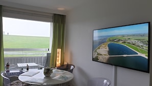 Hotelkamer met een groot raam, een tafel met een bloevas, een stoel en een flatscreen-tv waarop een luchtbeeld van een kustgebied te zien is.