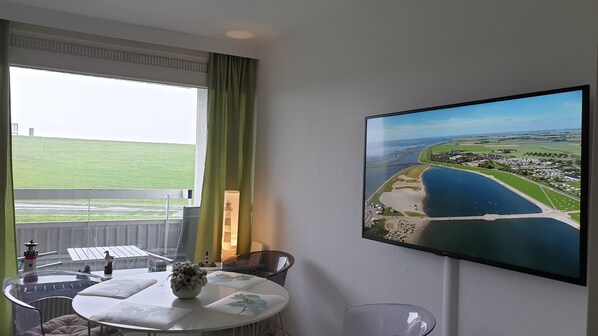 A hotel room with a large window, a table with a vase of flowers, a chair, and a flat-screen TV displaying an aerial view of a coastal area.