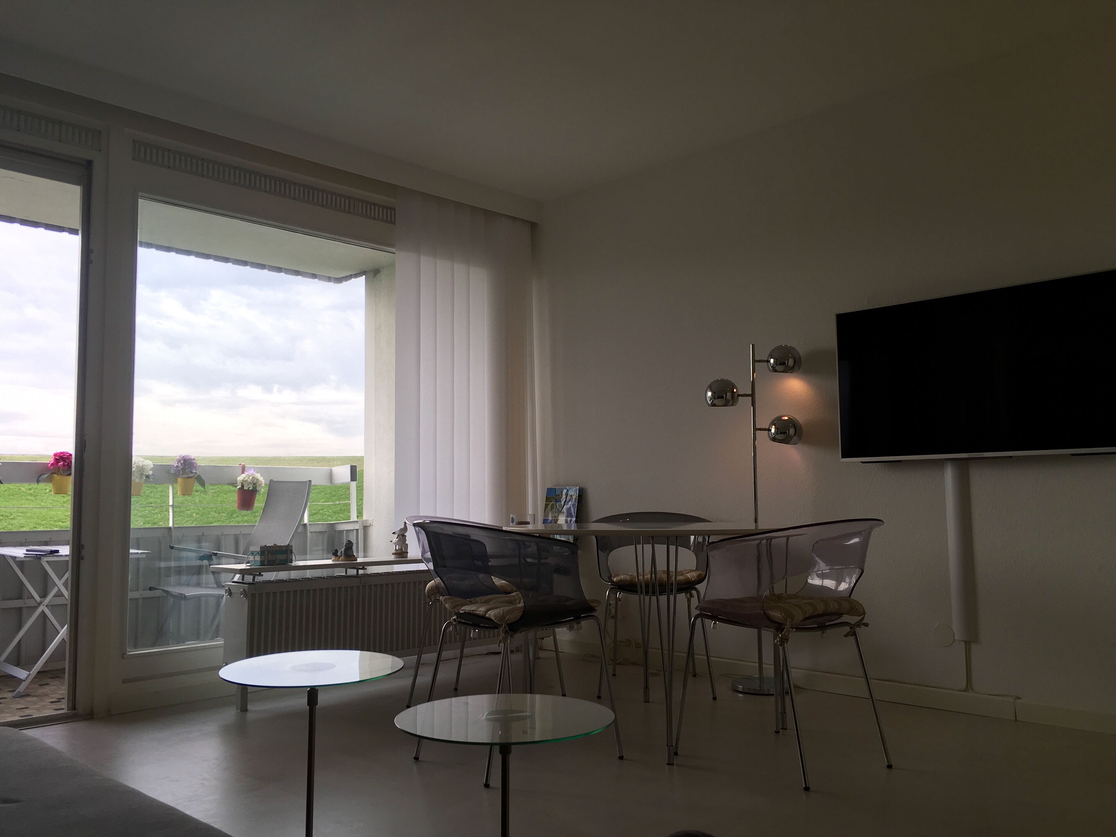 Ein modernes Wohnzimmer mit großem Fenster, einem wandmontierten Flachbildfernseher und einer Sitzecke mit zwei Tischen mit Glastischplatte sowie vier Stühlen.