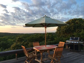 An outdoor deck with a table and chairs, a grill, and a view of a forested landscape. - Light Open Vacation Home, Sweeping Waterviews, Hill Top, Private  (Chilmark)