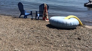 Ein Kiesstrand mit zwei blauen Stühlen, einem überdachten Picknicktisch, einem gelben aufblasbaren Schwimmreifen und einem Ruderboot.