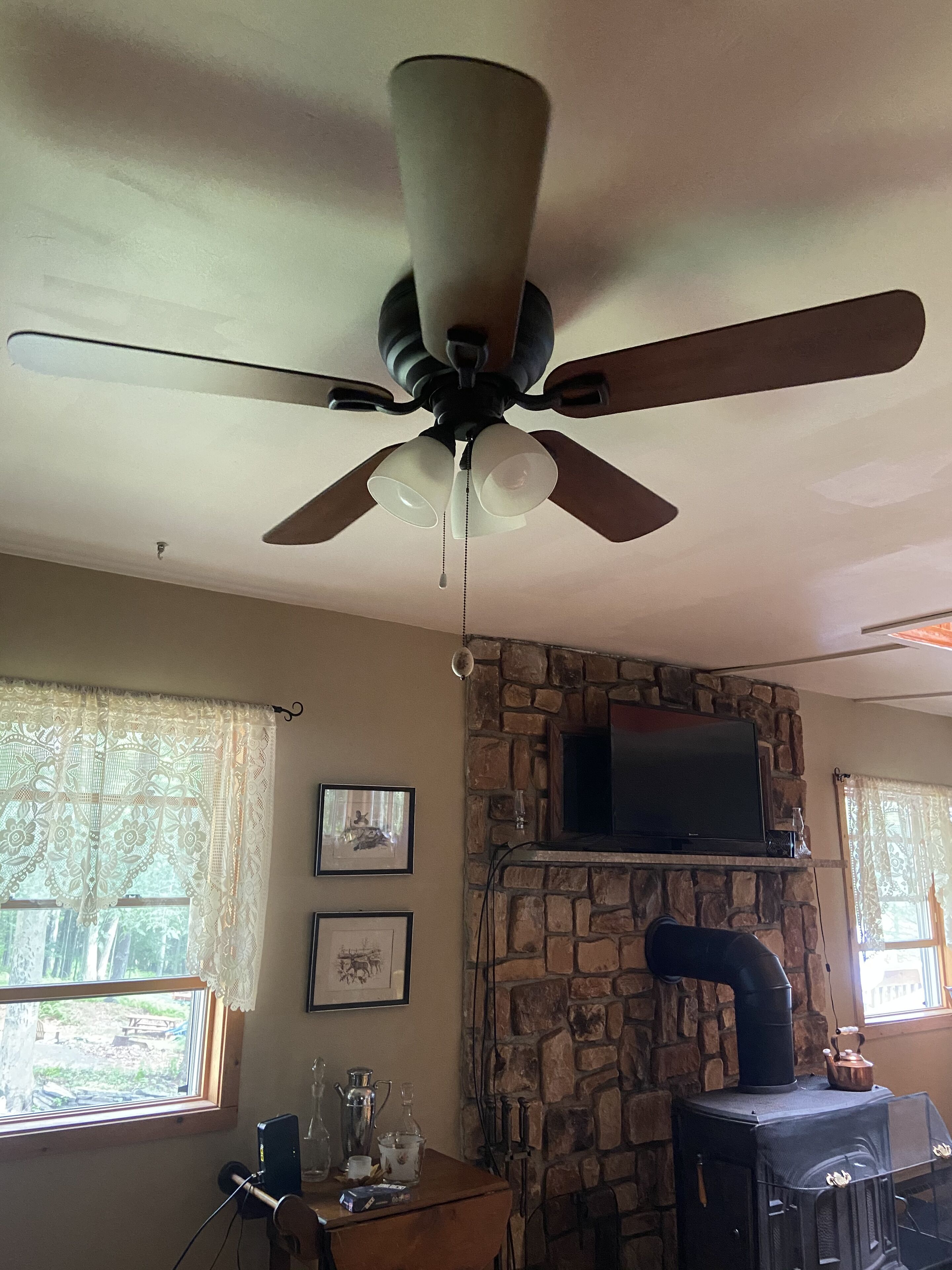 Une pièce dotée d’un ventilateur de plafond, d’une cheminée en pierre, d’un téléviseur fixé au-dessus de la cheminée et d’une fenêtre ornée de rideaux en dentelle.