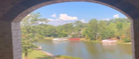 A view through an arched window of a lake surrounded by trees and a grassy area.