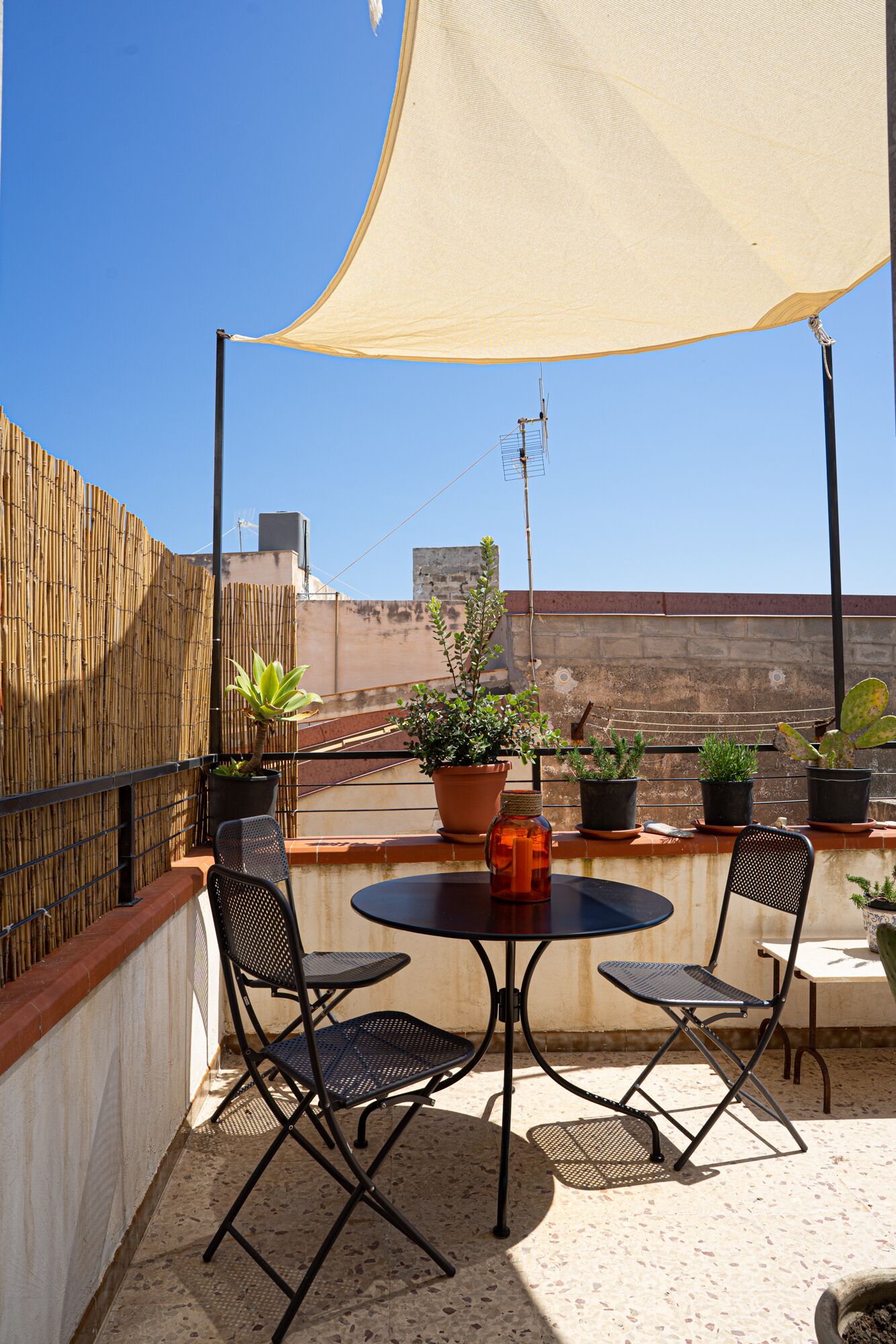 Een terras op het dak met een ronde tafel en twee stoelen, omringd door potplanten en een grote parasol.