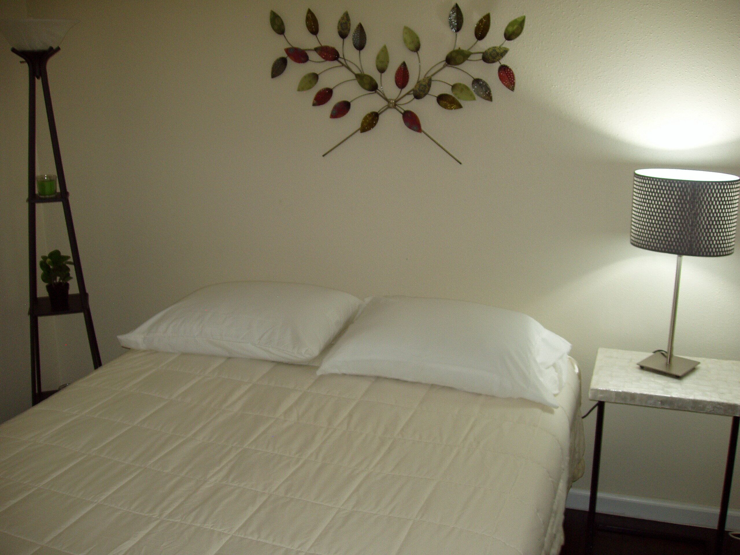 En seng med hvide sengetøj, et vægmaleri med bladmotiver, et lille bord med en lampe og en stige med en potteplante.