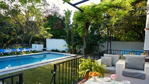 Un espace aménagé au bord de la piscine, avec un canapé blanc pour l’extérieur, une table en verre surmontée d’assiettes blanches et d’un vase rempli de fleurs, et une piscine bleue.