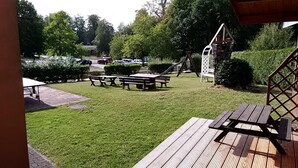 Un espace extérieur verdoyant avec des tables de pique-nique, une aire de jeux et un parking en arrière-plan.