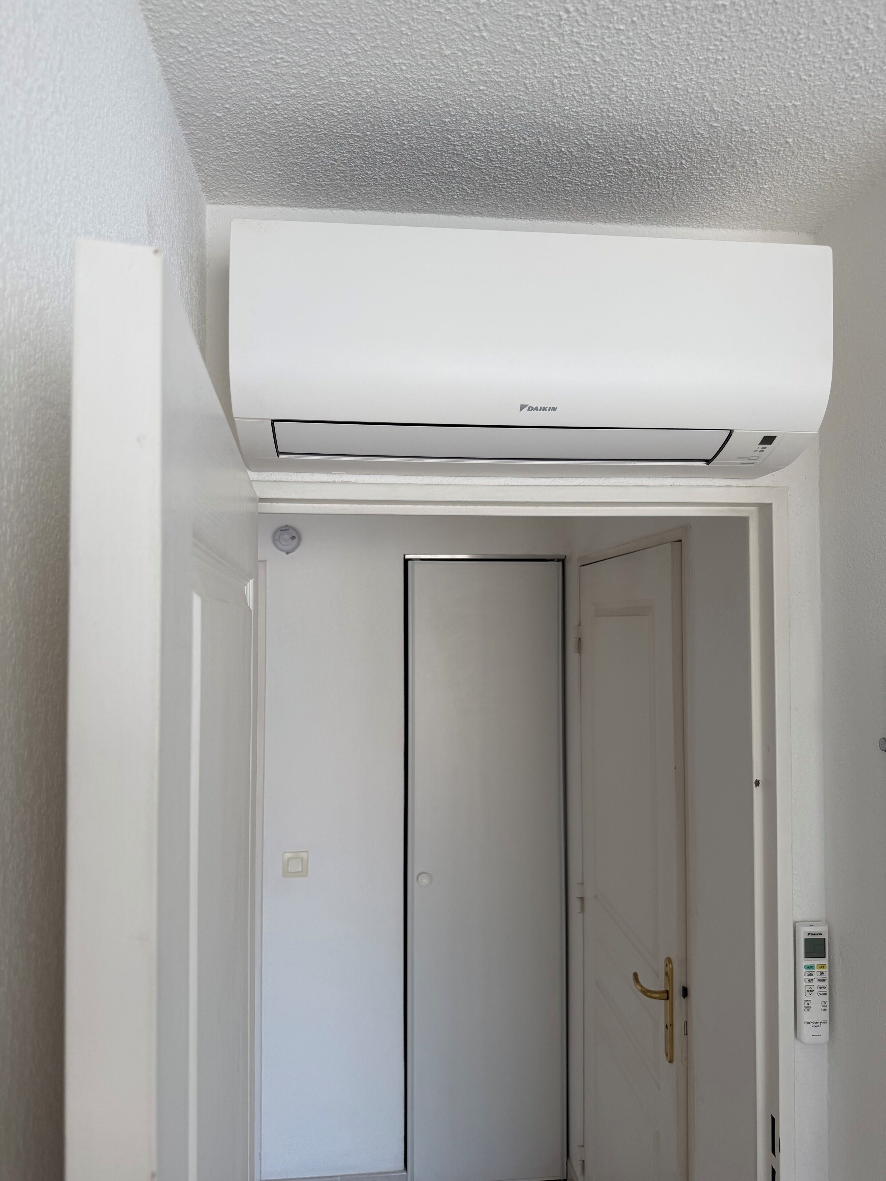 Un equipo de aire acondicionado blanco montado en el techo, una puerta blanca con pomo dorado y un panel de control fijado a la pared.