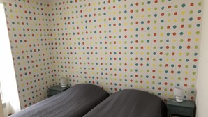 Quarto com duas camas, papel de parede estampado com bolinhas e uma pequena mesa de cabeceira com abajur.